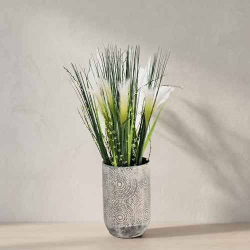 Artificial pampas grass arrangement in a textured gray ceramic pot, set against a neutral beige wall and light wood surface.
