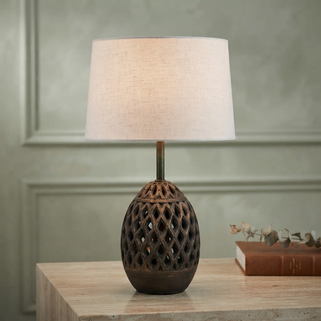A table lamp with a textured, dark brown lattice-patterned ceramic base and a light beige linen drum shade, shown lit on a light wood table against a muted green paneled wall.
