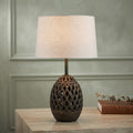A table lamp with a textured, dark brown lattice-patterned ceramic base and a light beige linen drum shade, shown lit on a light wood table against a muted green paneled wall.