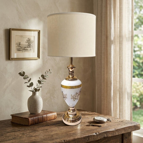 An ornate table lamp with a white ceramic body decorated with gold Greek key and floral motifs, topped with a gold finial and a beige fabric drum shade, styled on a rustic wooden table in a warm, sunlit room.