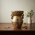 A rustic bronze-toned ceramic vase with two side handles and a decorative key motif on the front, displayed on a wooden table with a white pitcher of flowers in the background.