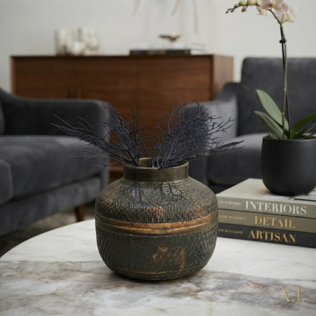 A dark textured ceramic vase with a distressed bronze-brown band, holding dried dark branches, styled on a marble table in a modern living room setting.