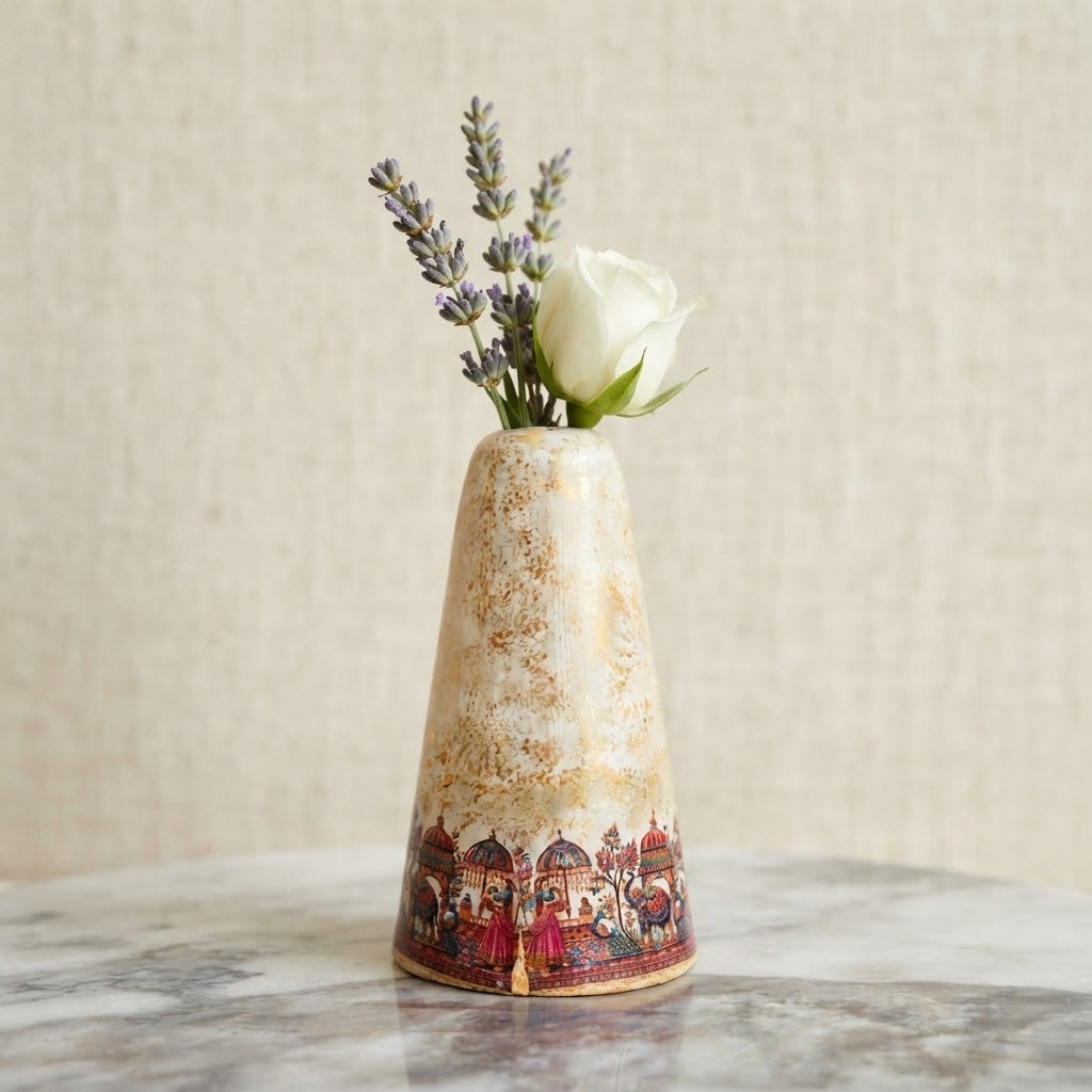 A small conical ceramic vase with a distressed cream and gold finish and a red illustrated border near the base, holding a white rose and lavender stems on a marble surface against a neutral textured wall.