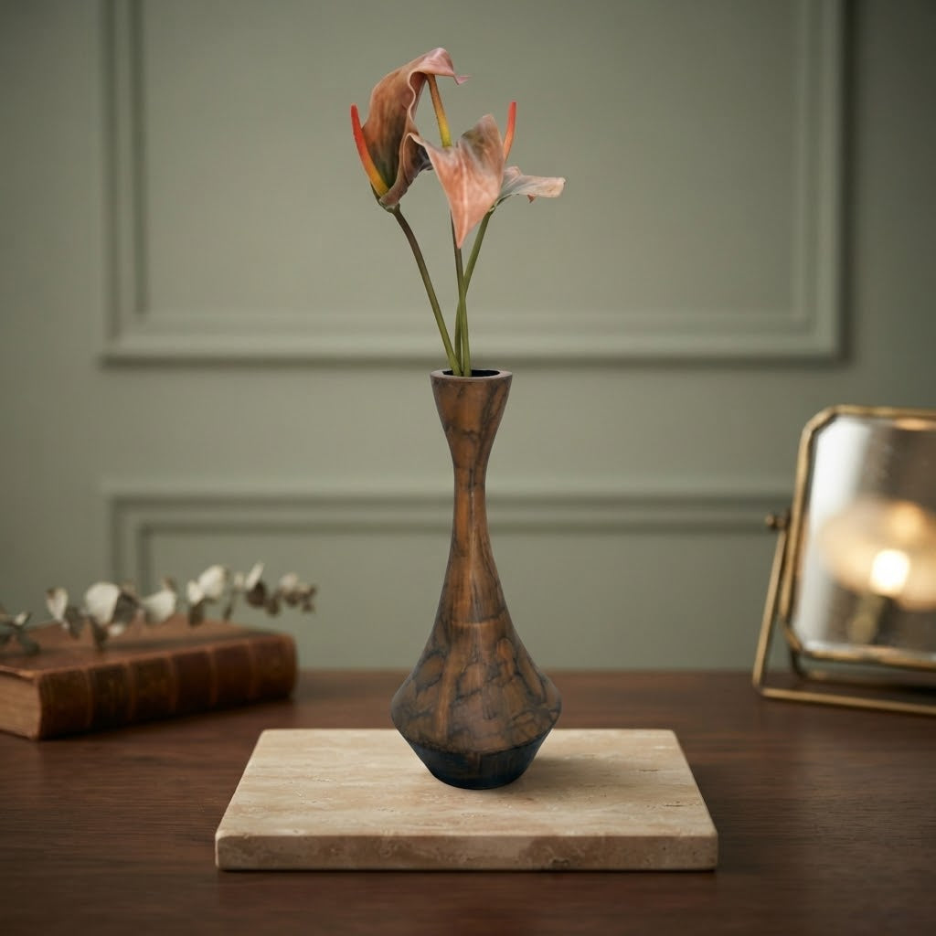 A small sculptural ceramic vase with a dark brown and black marbled pattern, holding two dried pinkish-brown flowers, displayed on a light stone tray on a wooden table.
