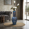 A tall blue ceramic vase with embossed floral details stands on a round woven rug in a styled living room. White flowers are placed in the vase, and the room includes a gray tufted sofa, a small wooden side table, and a window with curtains.
