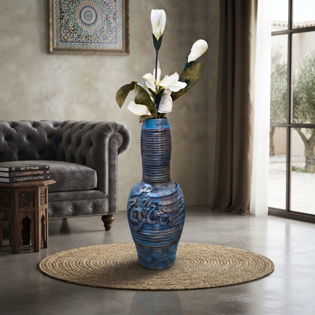 A tall blue ceramic vase with embossed floral details stands on a round woven rug in a styled living room. White flowers are placed in the vase, and the room includes a gray tufted sofa, a small wooden side table, and a window with curtains.
