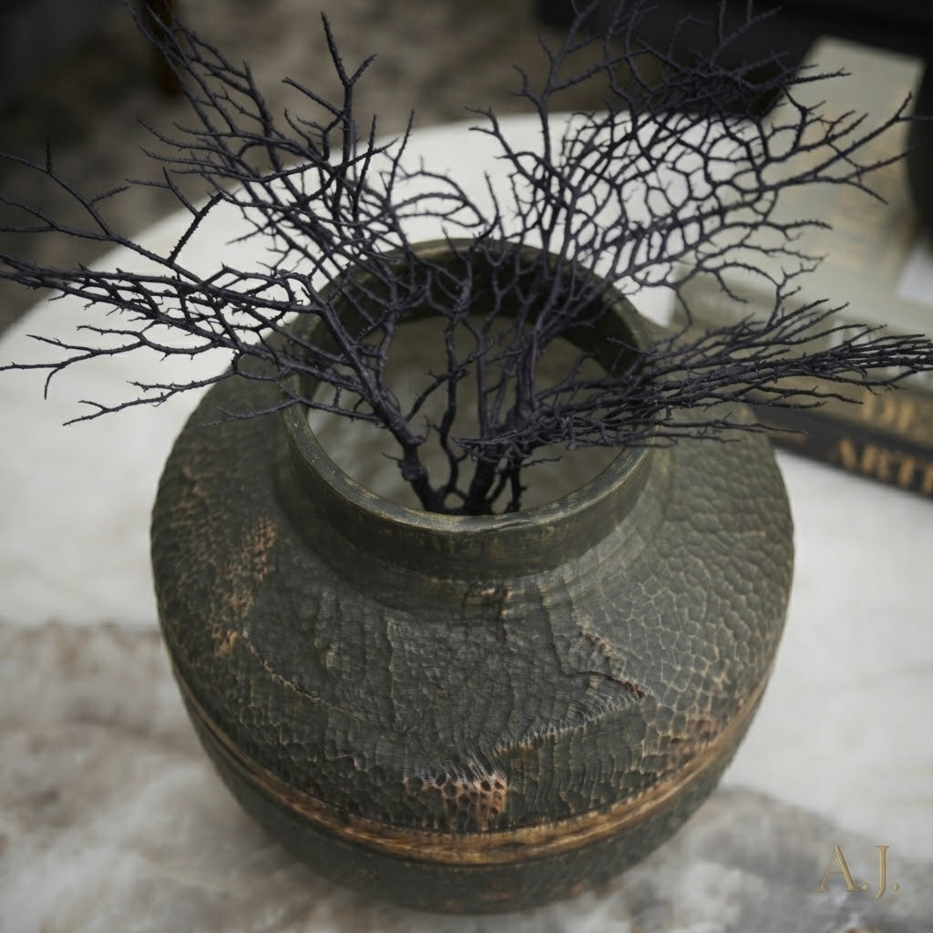 A dark textured ceramic vase with a hammered finish, holding dried black coral branches, set on a light marble surface.
