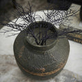 A dark textured ceramic vase with a hammered finish, holding dried black coral branches, set on a light marble surface.